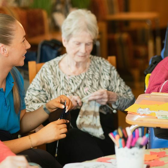 Pflegekraft im Gespräch mit zwei Seniorinnen bei einer gemeinsamen Handarbeit