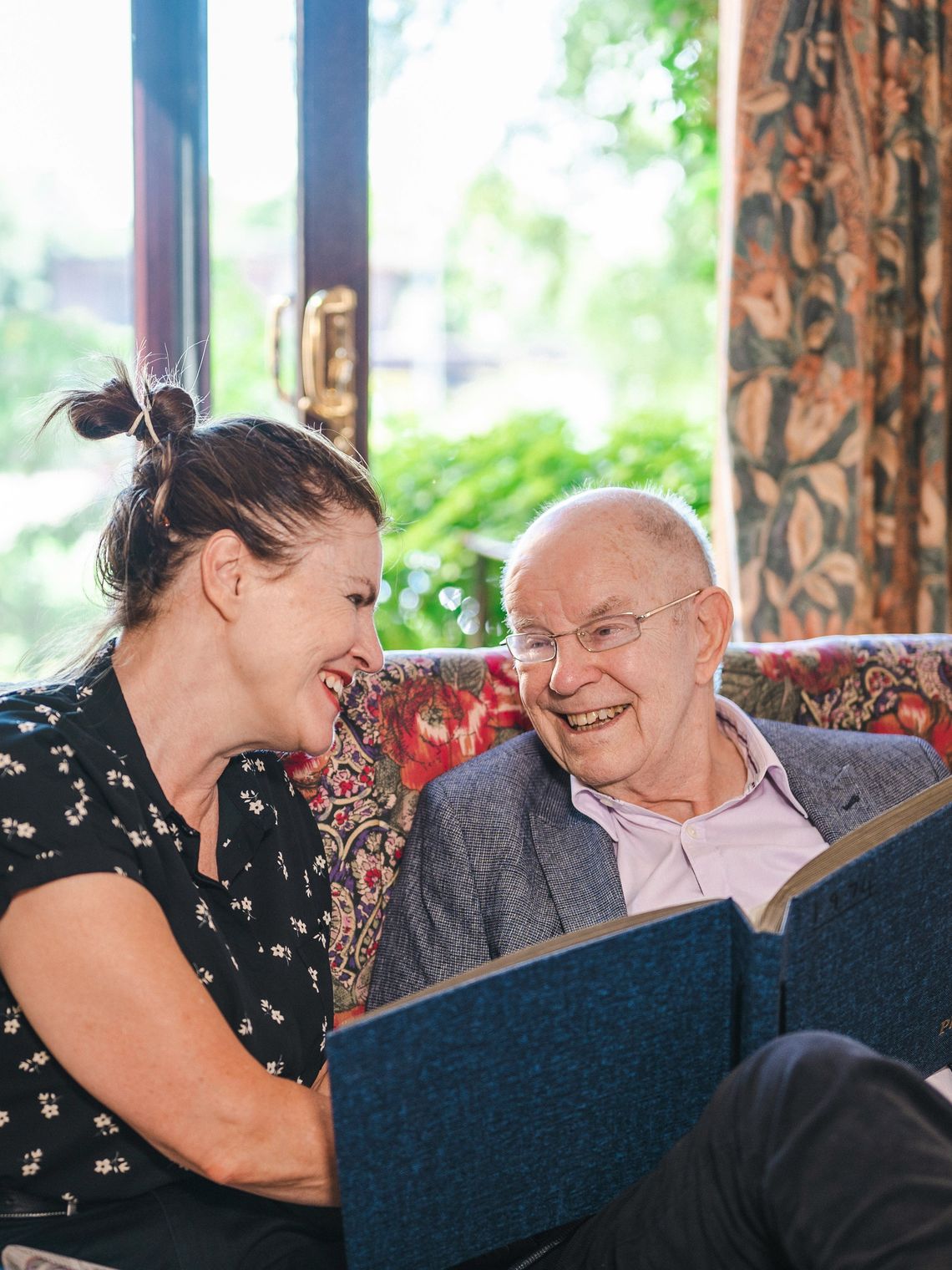 Pflegekraft und älterer Herr sitzen lachend auf dem Sofa und blättern gemeinsam in einem Fotoalbum